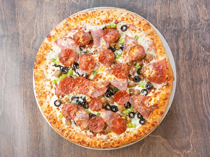 A whole pizza with pepperoni, ham, sausage, green peppers, and black olives on a wooden table.