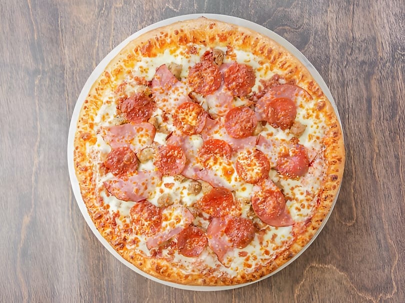 A whole meat lover's pizza with pepperoni, ham, and sausage on a wooden table.