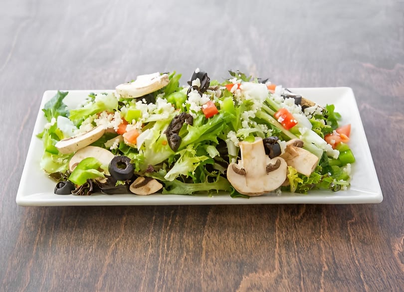 Fresh garden salad with lettuce, tomatoes, mushrooms, olives, and crumbled cheese on a white rectangular plate.