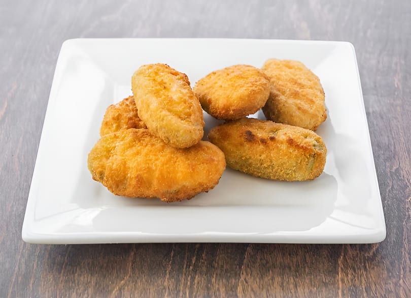 A white rectangular plate holds a pile of golden-brown, breaded jalapeño poppers on a wooden surface.